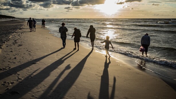 Une scène de plage où l’on voit des personnes se promener le long du rivage, et où l’on distingue les ombres de ces personnes et du soleil sur le sable.
