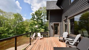 Une terrasse en bois avec des sièges extérieurs et une balustrade en verre donnant sur une zone boisée.