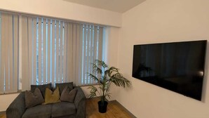 Ein Wohnzimmer mit grauem Sofa, einem Topf mit Pflanze und einem großen Flachbildfernseher an der Wand.