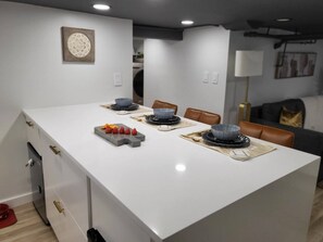 A modern kitchen with a central island, white cabinetry, and a dining area with chairs and a table set for a meal. - Romantic hideout. 1Br. Lower Level (Stamford)