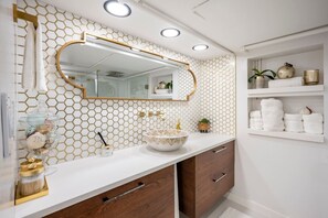 Un bagno con rivestimento a mosaico esagonale bianco e dorato, uno specchio tondo, un lavandino bianco con una ciotola dorata e uno scaffale con articoli da toilette.