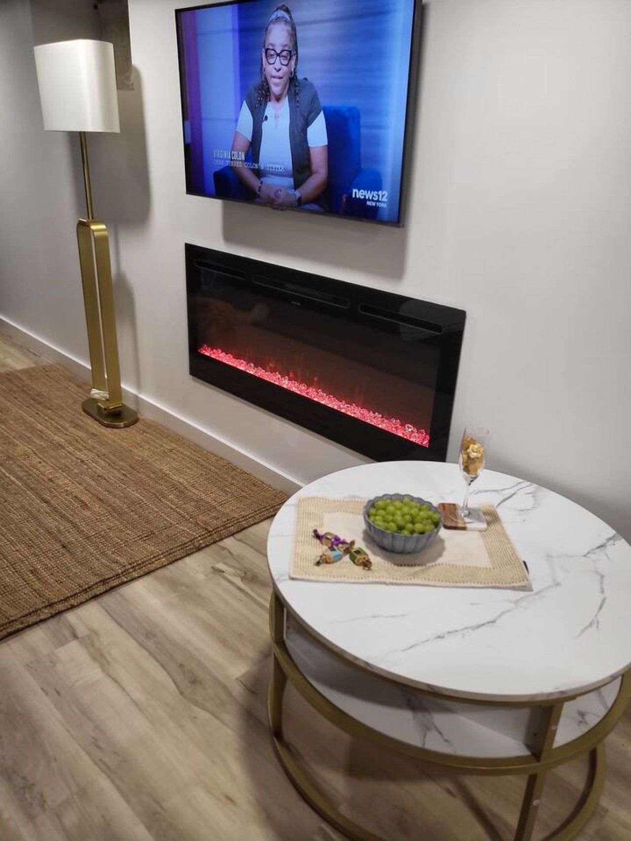 Ein modernes Wohnzimmer mit einem Flachbildfernseher, einem Kamin, einem runden Marmortisch mit einer Schale Trauben und einer Stehlampe.
