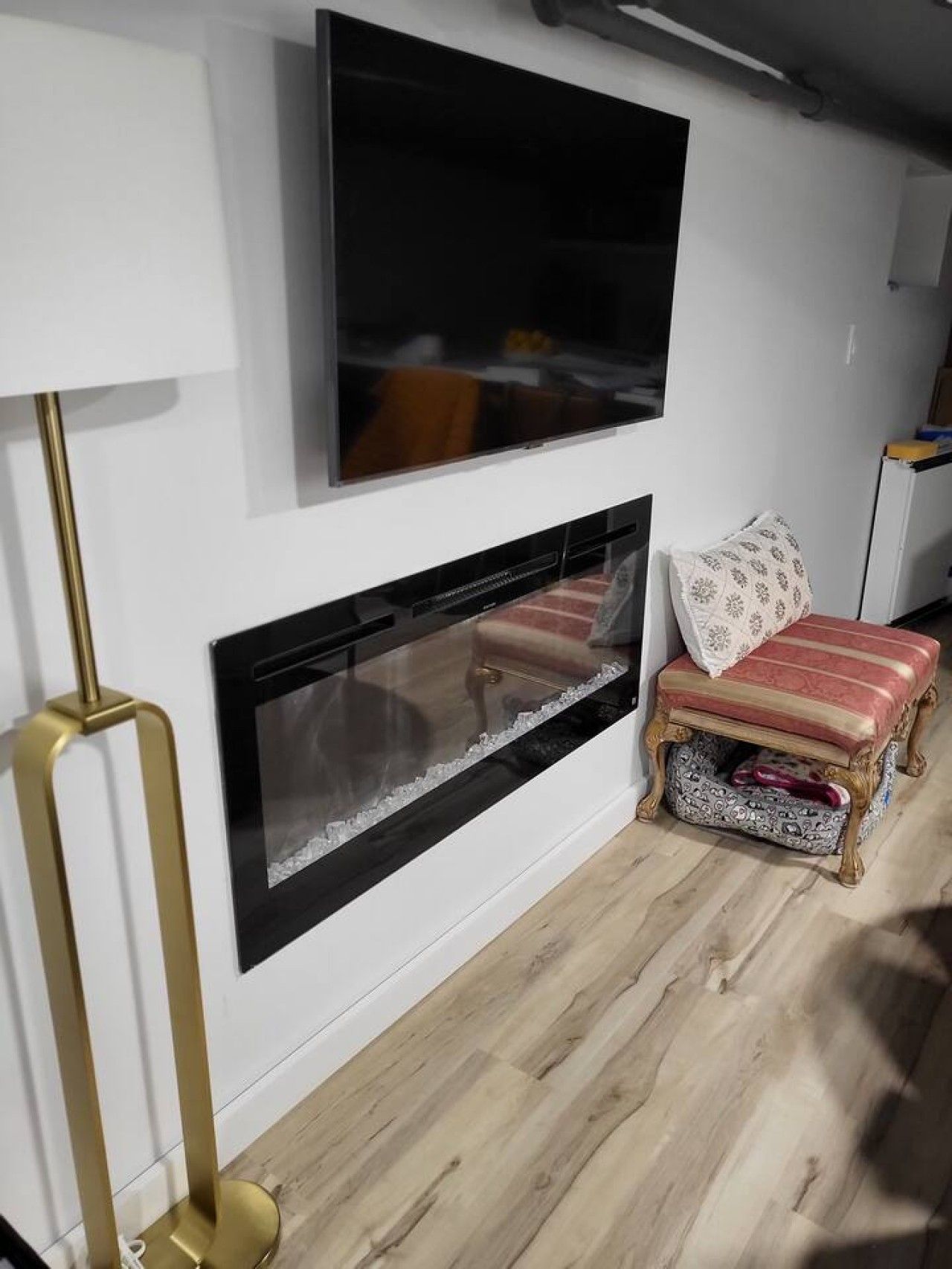 Ein Zimmer mit einem flachbildfernseher über einem Kamin, einer Stehlampe und einer gepolsterten Bank mit gemustertem Kissen.