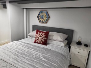 A bedroom with a bed, a nightstand, a decorative wall piece, and a small table with a lamp. - Romantic hideout. 1Br. Lower Level (Stamford)
