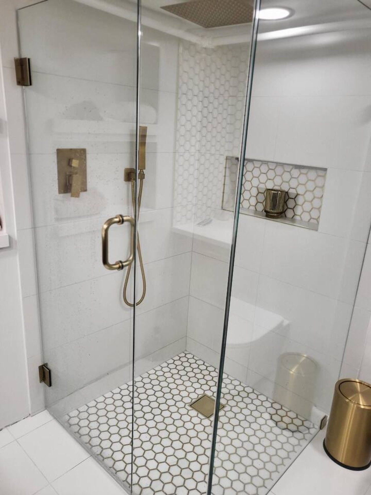 Una ducha con cerramiento de vidrio, suelo de baldosas hexagonales y ducha de color dorado.