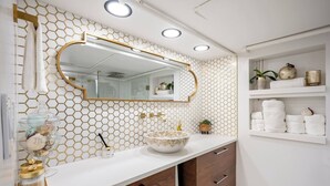 A bathroom with a white vanity, a gold-framed mirror, a white hexagonal tile backsplash, and a shelf with toiletries. - Romantic Hide Out
 (Stamford)