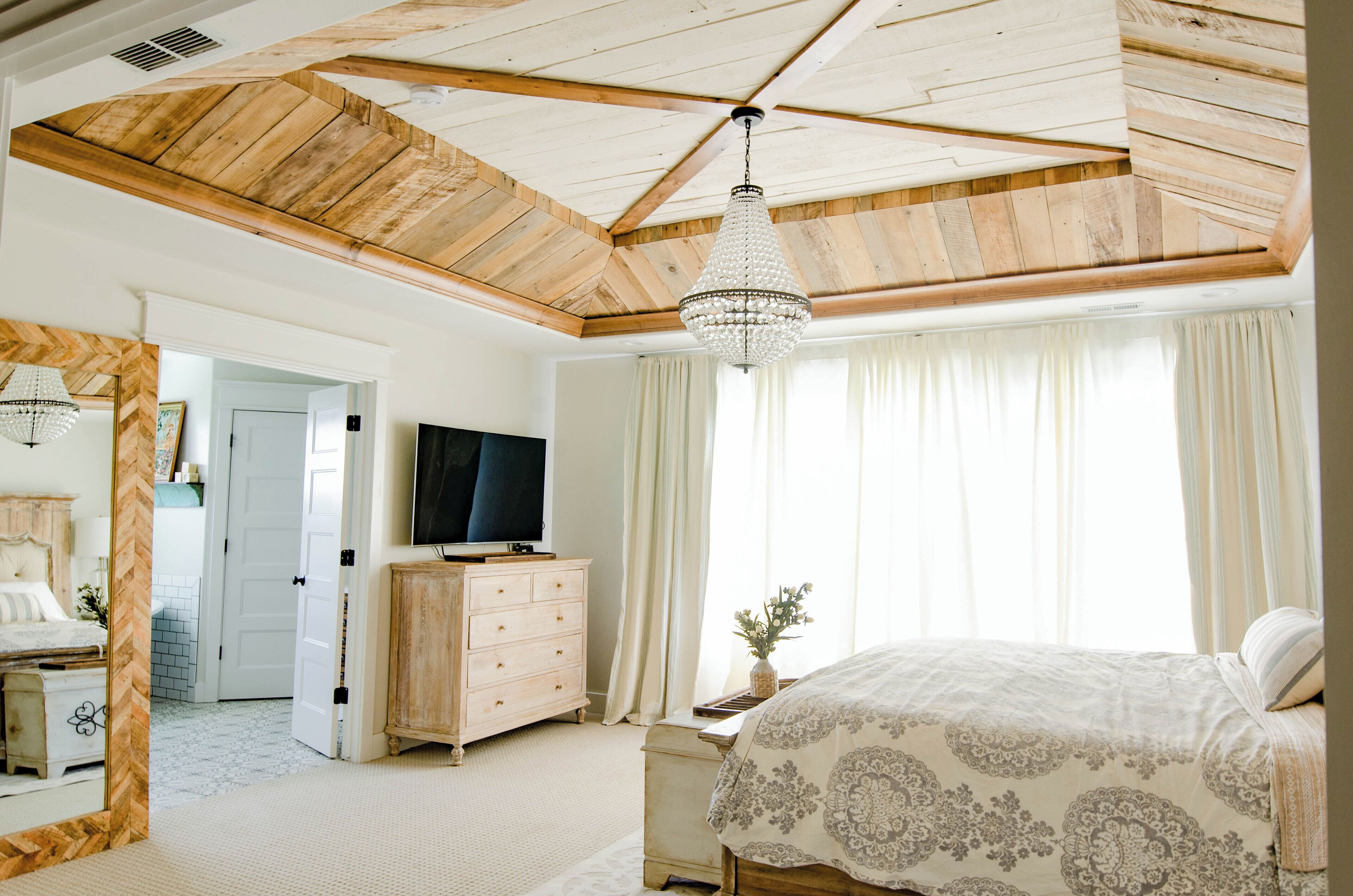 A bedroom with a wooden ceiling, a chandelier, a bed with a patterned bedspread, a dresser, and a television.
