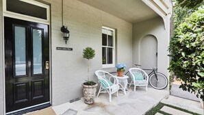 A house entrance with a black door, a white wall, a potted tree, a bicycle, and outdoor seating with chairs and a table.