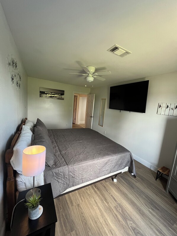 A bedroom with a bed, a bedside lamp, a ceiling fan, a TV mounted on the wall, and a small wooden stool.