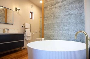 A bathroom with a round white bathtub, a dark wooden vanity, a large mirror, and a stone wall. - The Verdict Suite (Martinborough)