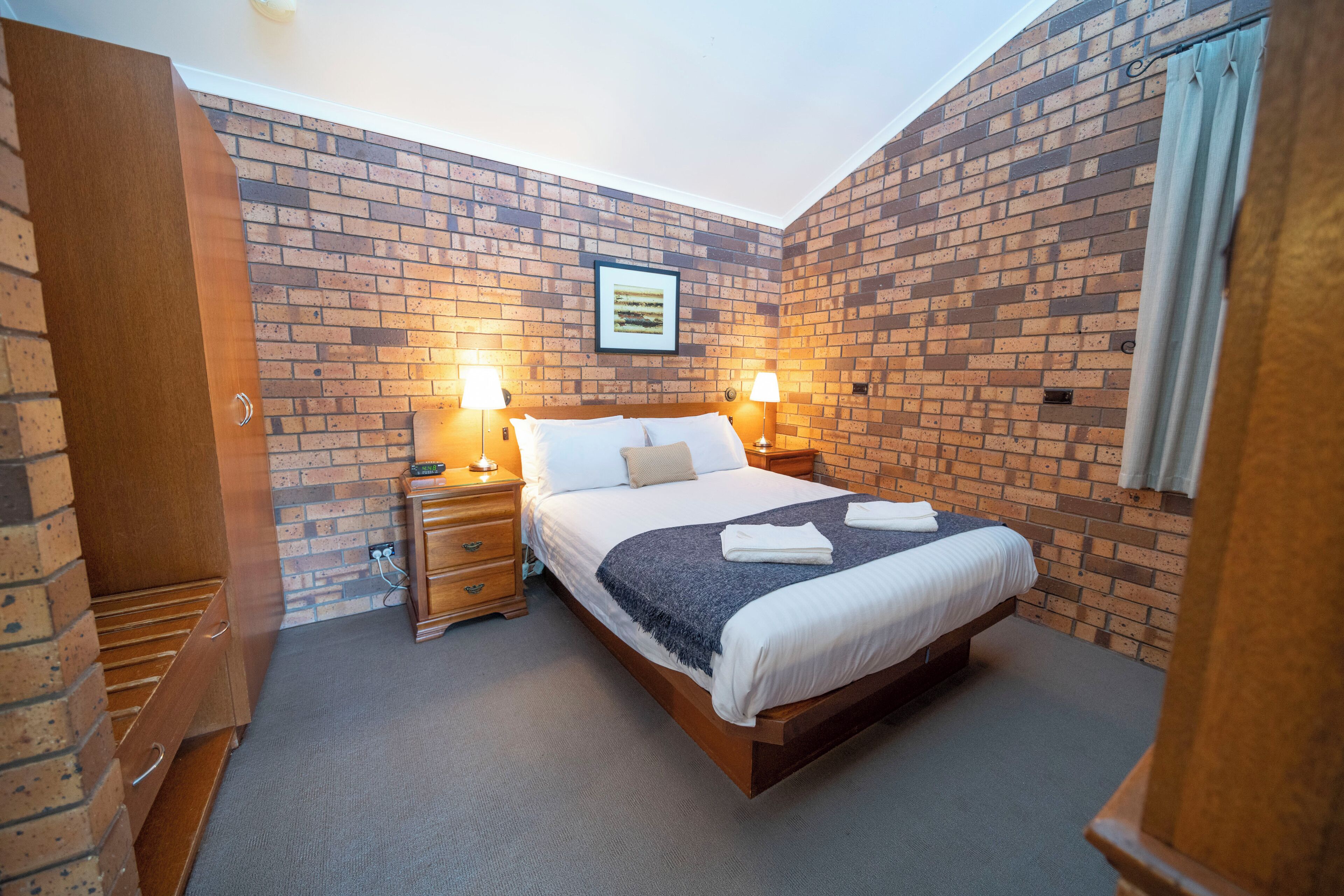Habitación de hotel con pared de ladrillo, una cama con ropa blanca, dos lámparas de noche y un cuadro enmarcado en la pared.