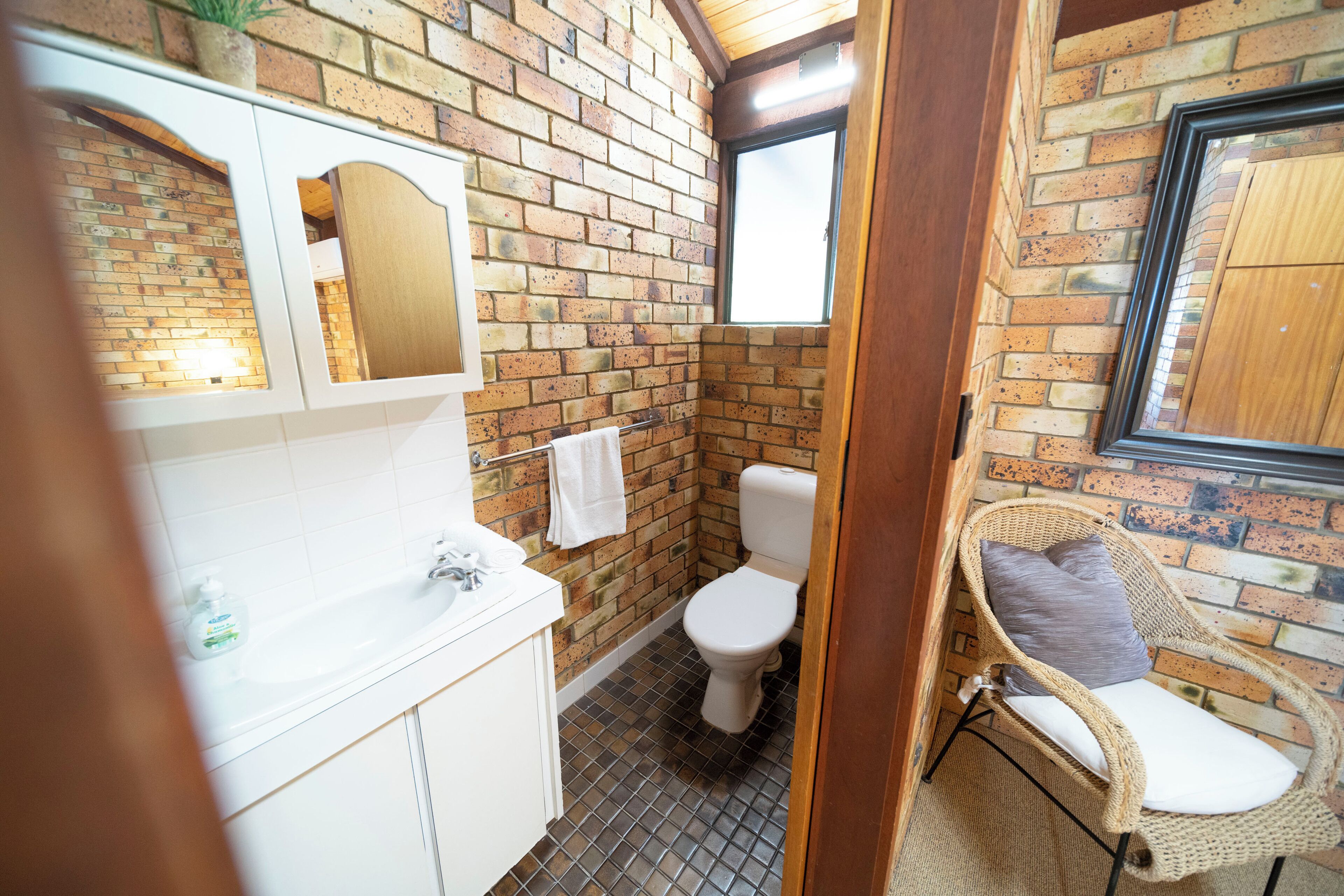 A bathroom with a brick wall, a white sink, a toilet, and a wicker chair.