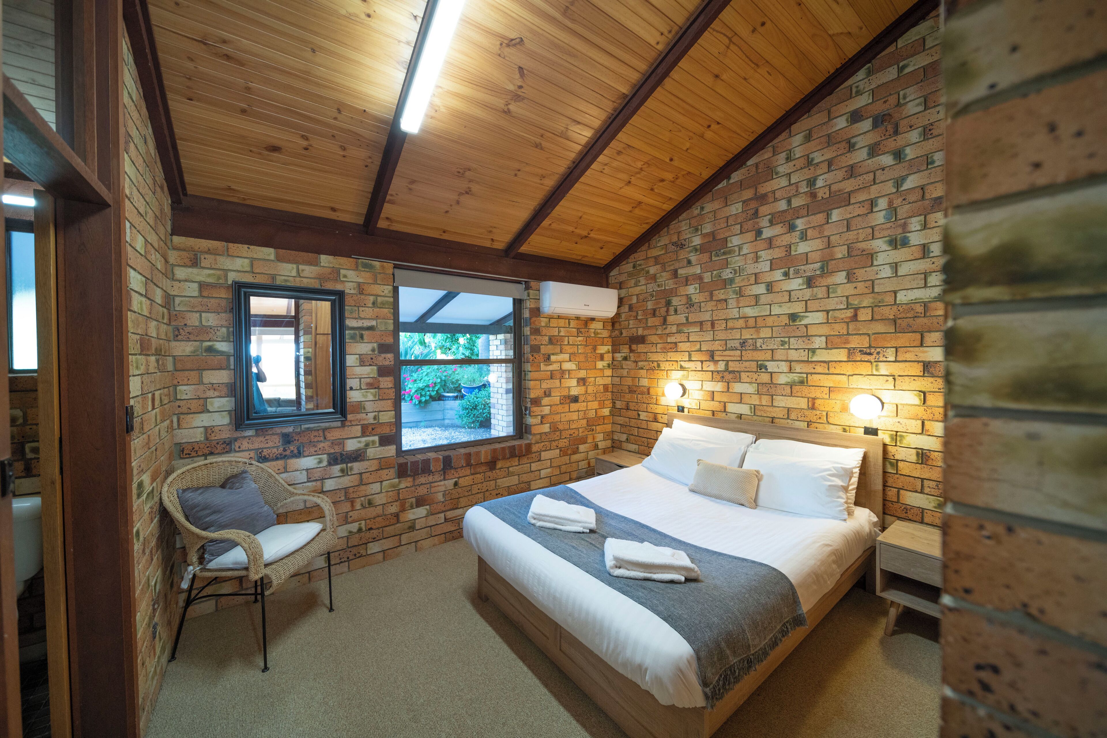 Una habitación con pared de ladrillo, vigas de madera en el techo, una cama con sábanas blancas, una silla de mimbre y una ventana con vista a la vegetación.