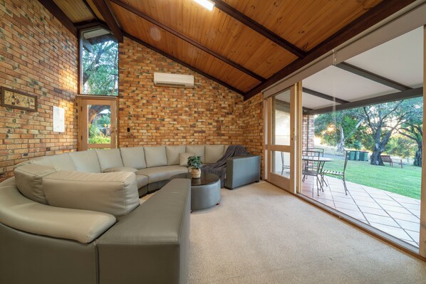 A spacious living area with a large sectional sofa, a round coffee table, and a brick wall. There is a sliding glass door leading to an outdoor patio. - Blickinstal Holiday Retreat (Bethany)