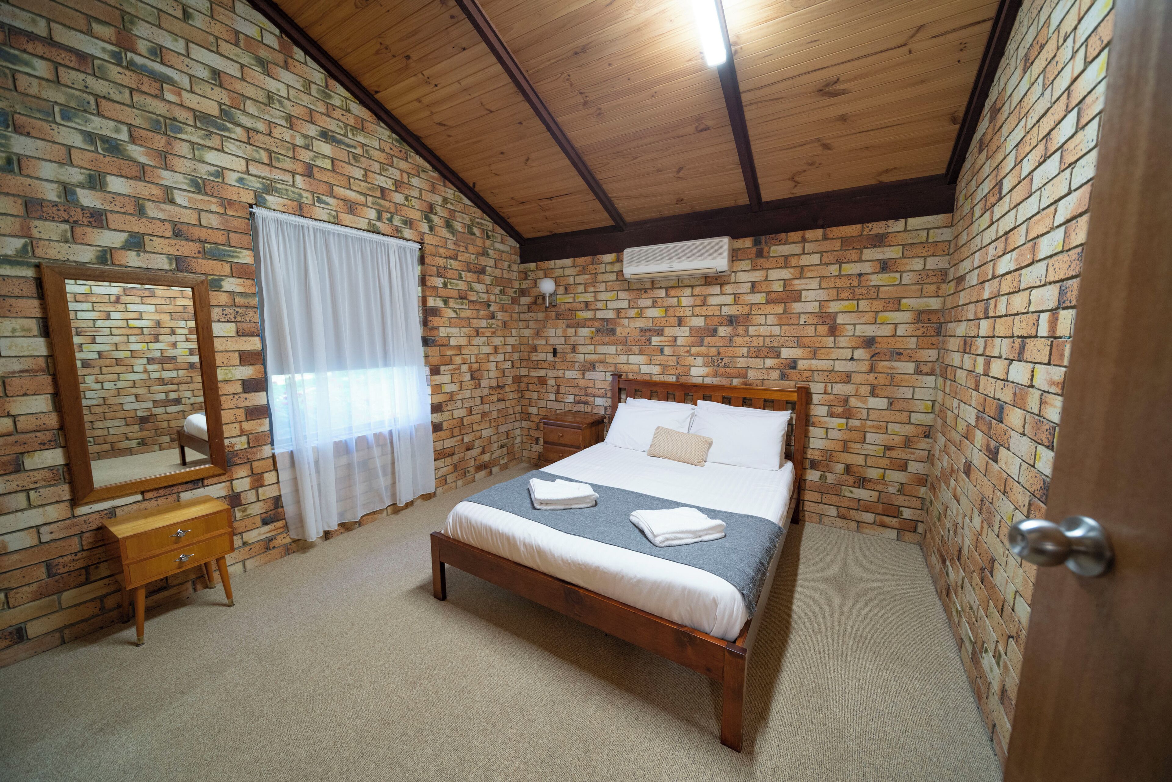 Un dormitorio con pared de ladrillo visto, vigas de madera en el techo, una cama de madera con ropa blanca, una mesita de noche de madera, un pequeño aparador de madera y un espejo en la pared.