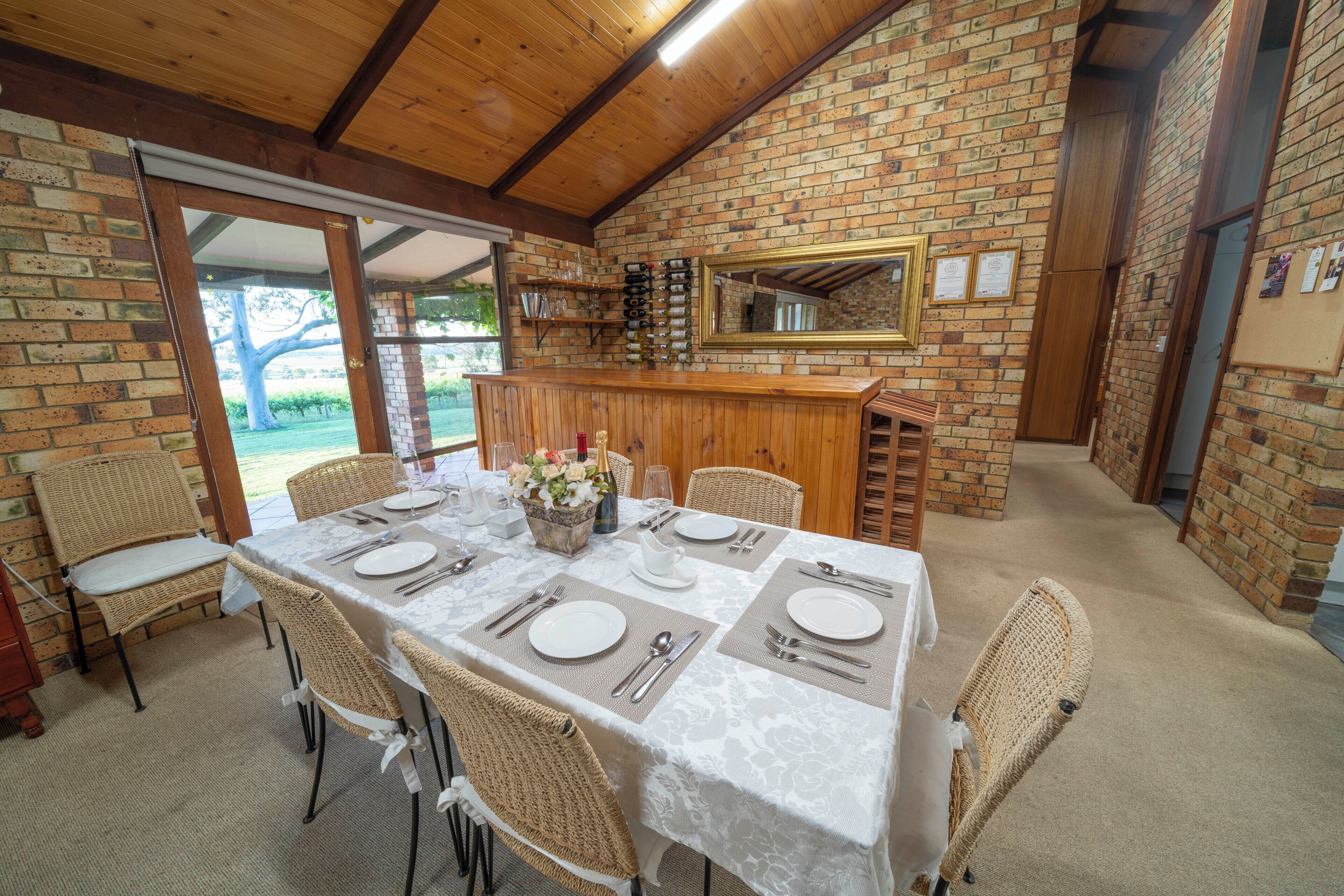 A dining area with a brick wall, wooden ceiling, and a table set for four with wicker chairs.