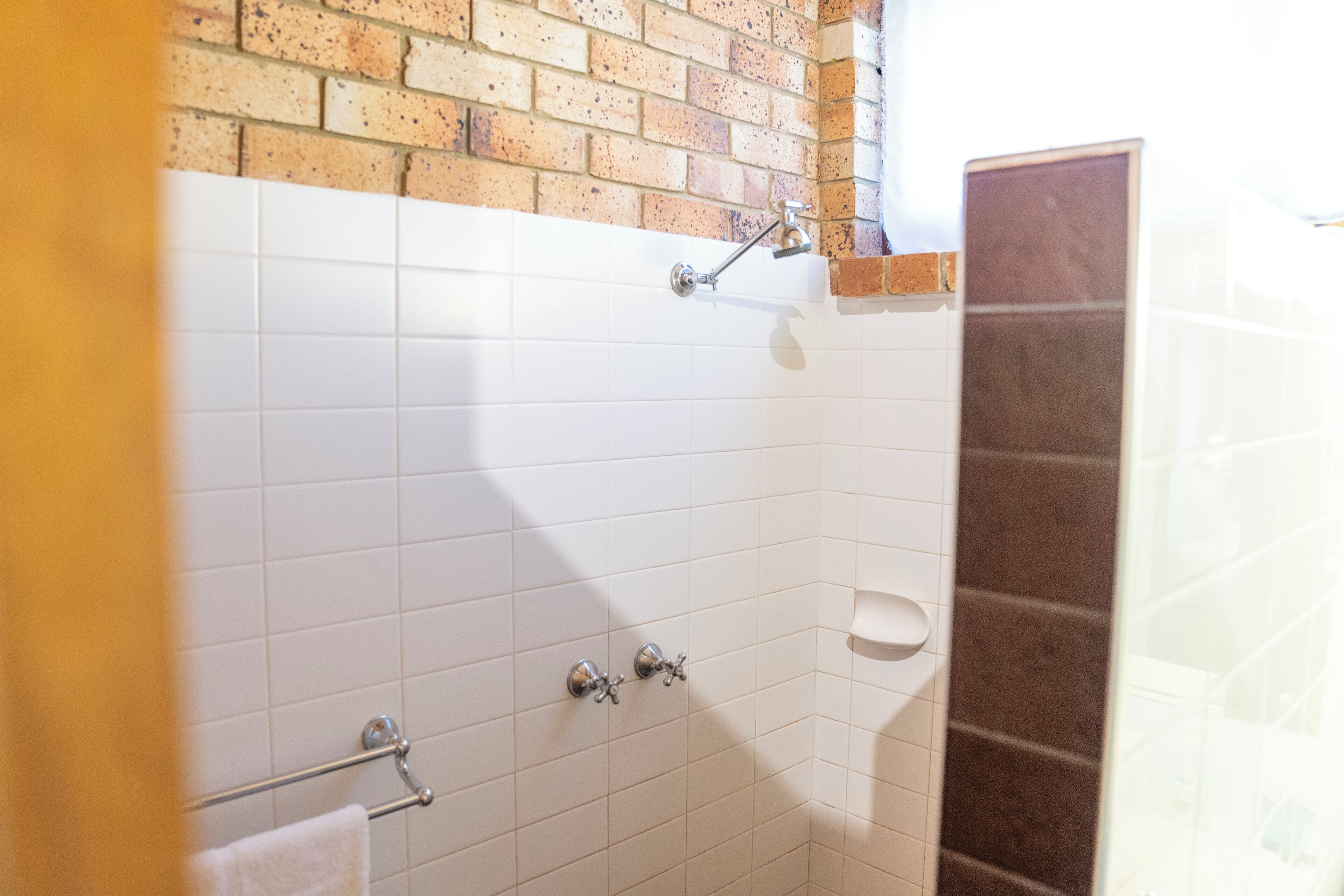 Un baño con paredes de azulejos blancos, una pared de acento de ladrillo, una ducha con cabezal de ducha de mano y un perchero.