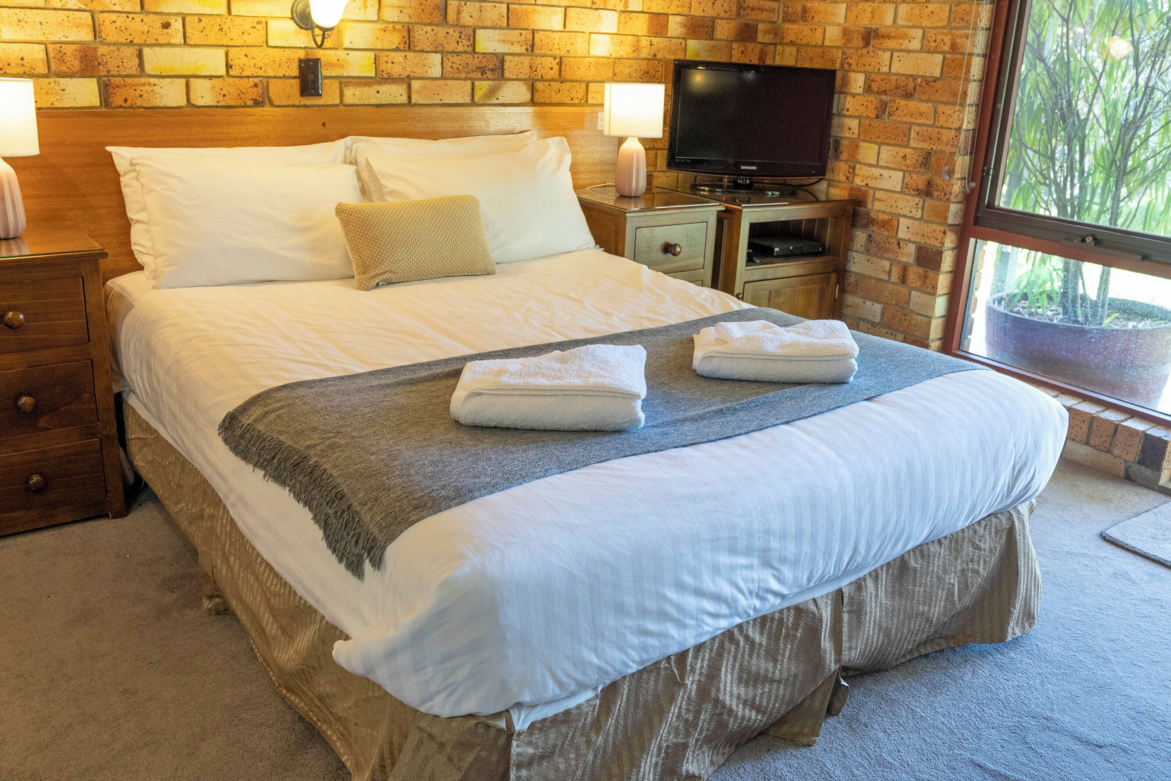 A neatly made bed with white linens, a grey throw blanket, and two folded towels. There is a television on a wooden stand beside the bed, and a brick wall with a window in the background.
