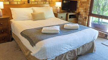A neatly made bed with white linens, a grey throw blanket, and two folded towels. There is a television on a wooden stand beside the bed, and a brick wall with a window in the background.