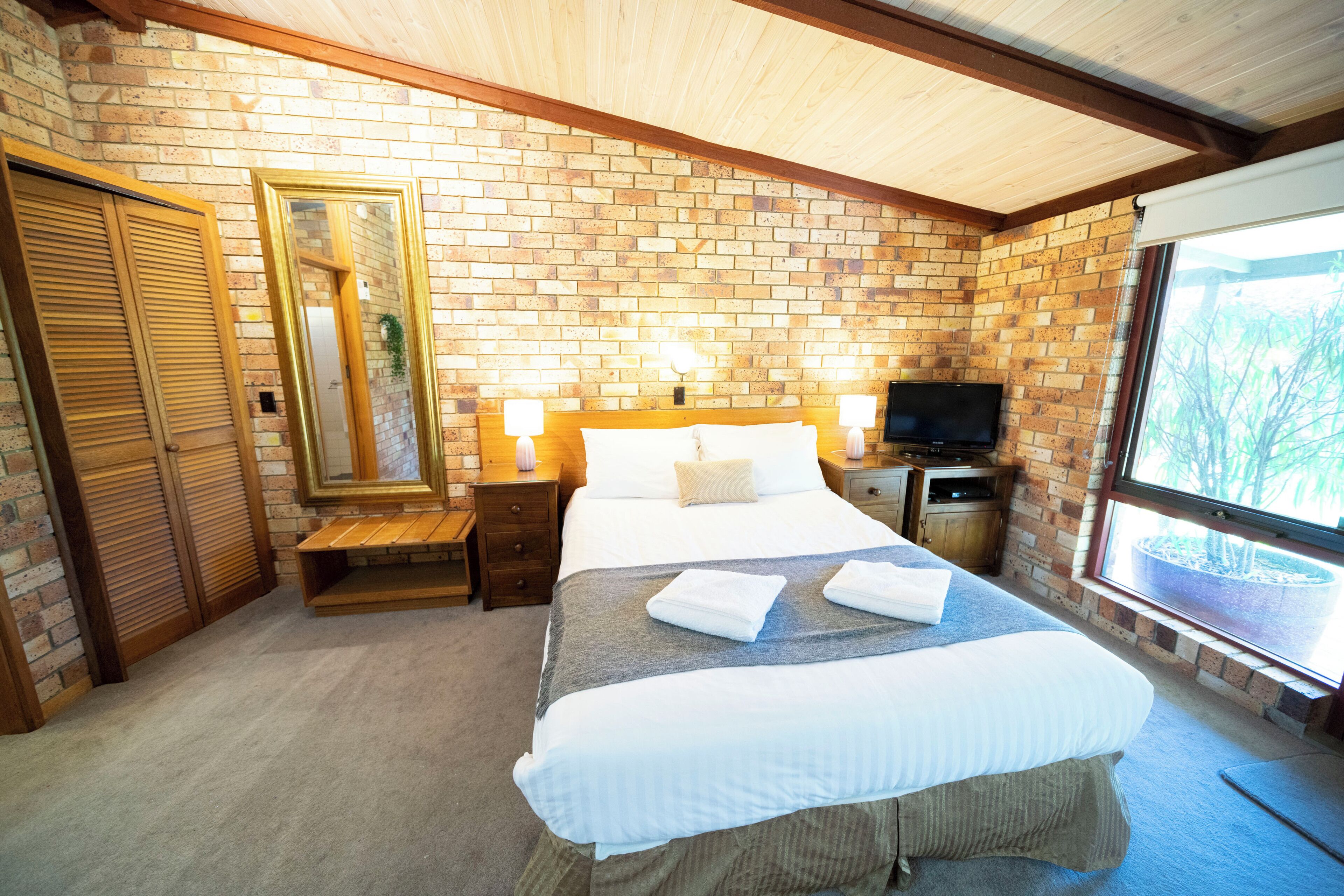 A bedroom with a brick wall, wooden ceiling beams, a bed with white linens, two bedside lamps, a TV on a wooden stand, and a window with a view of trees.