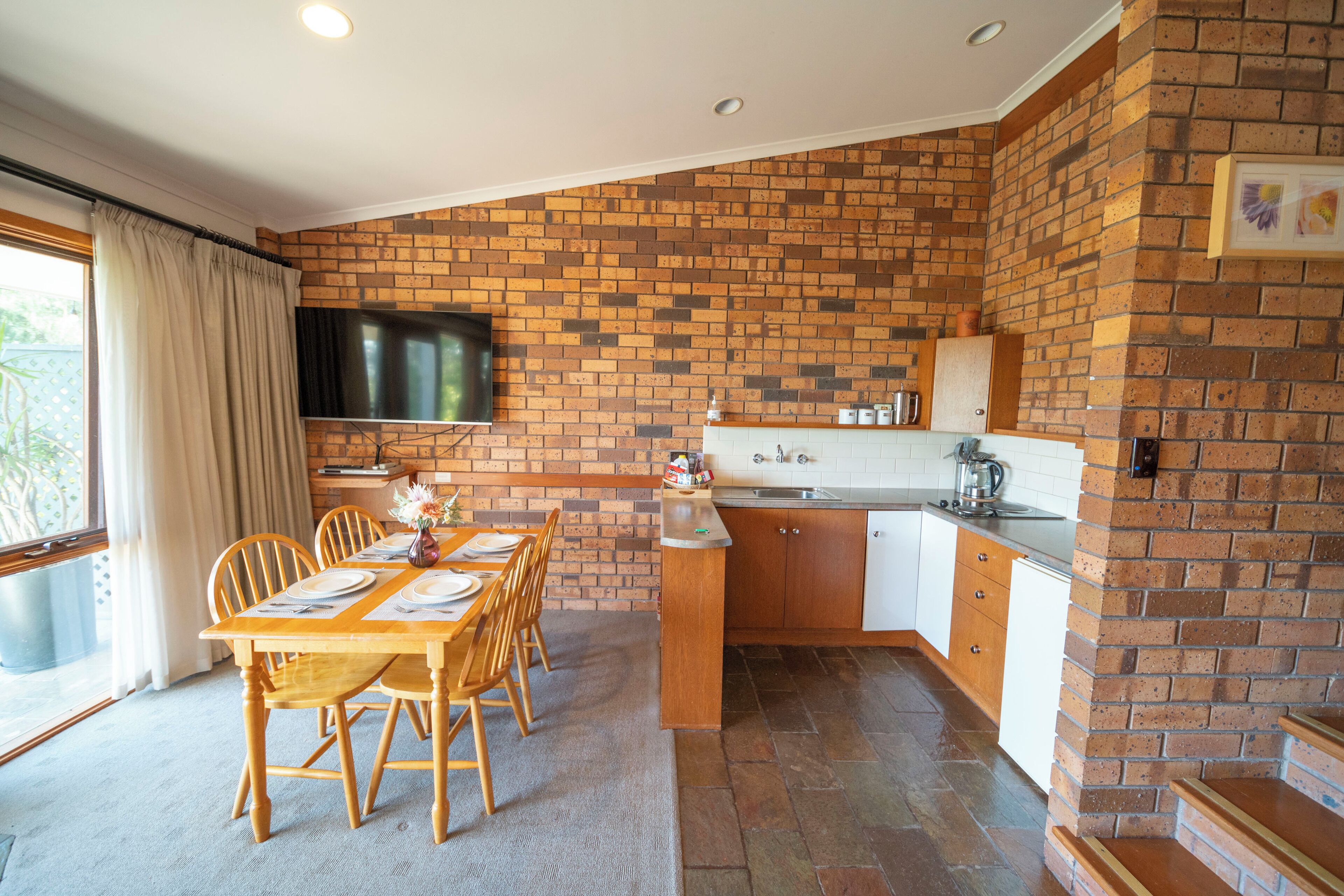 A dining area with a brick wall, a TV mounted above the counter, and a kitchen with white cabinets and a brick backsplash.