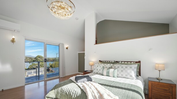 A spacious bedroom with a large bed, a nightstand, a chandelier, and a view through a sliding glass door. - Blossoms on the Bay - Nubeena (Nubeena)