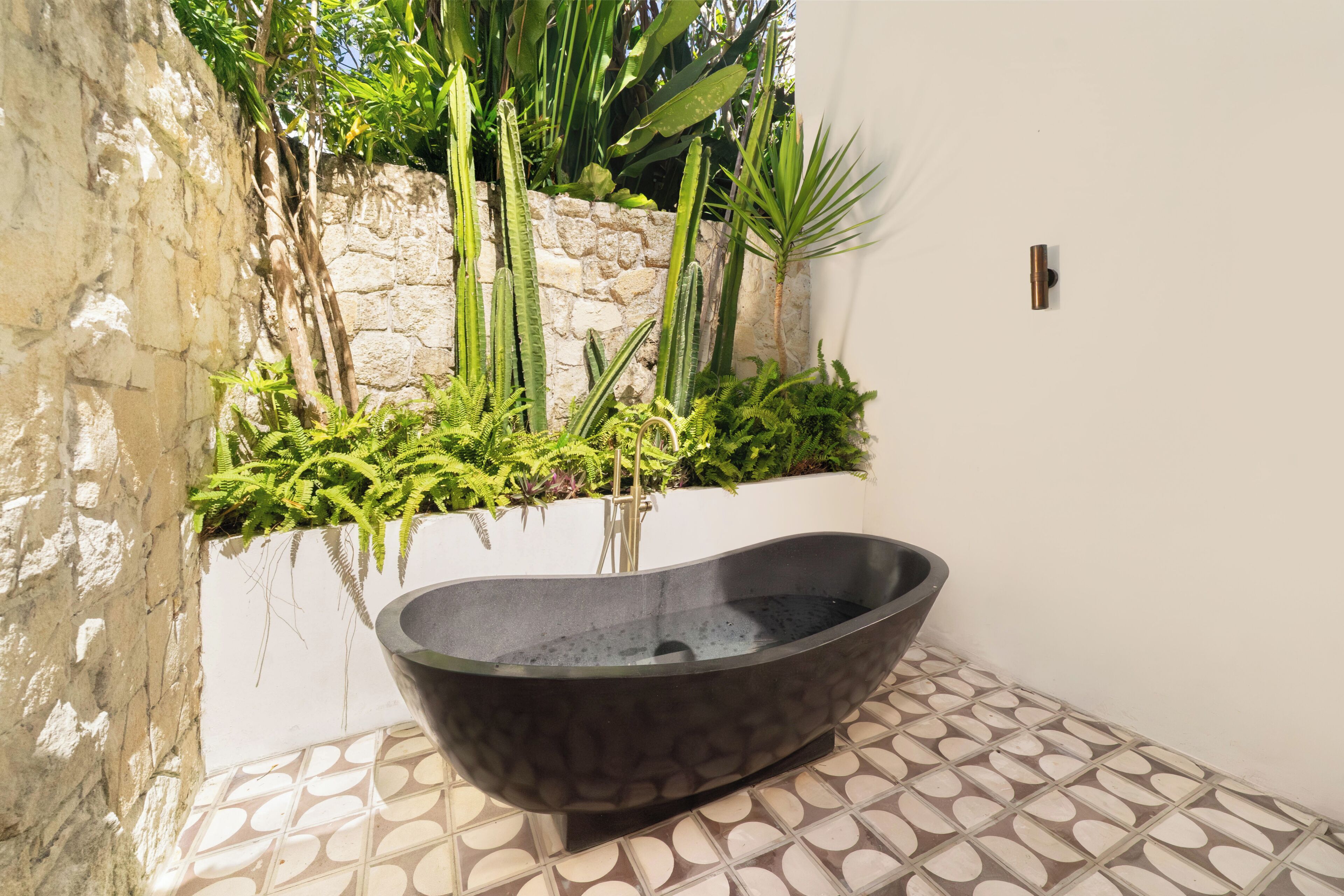 A stone bathtub in a tiled bathroom with a stone wall and greenery.