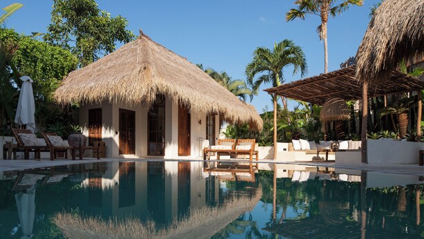 Zona de piscina con una estructura de techo de paja, sillas de descanso y una piscina cristalina que refleja el entorno.