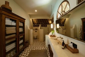 Un baño con suelo de baldosas, un mueble de madera con toallas, un lavabo con espejo y una ventana con vistas al tejado.