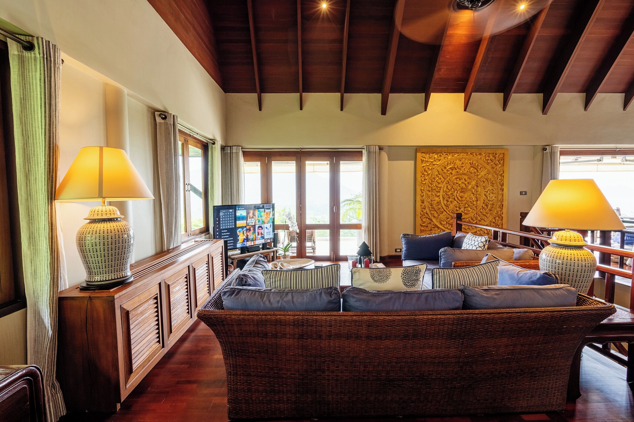 A spacious living area with a large wicker sofa, two lamps, a television, and a decorative wall panel.