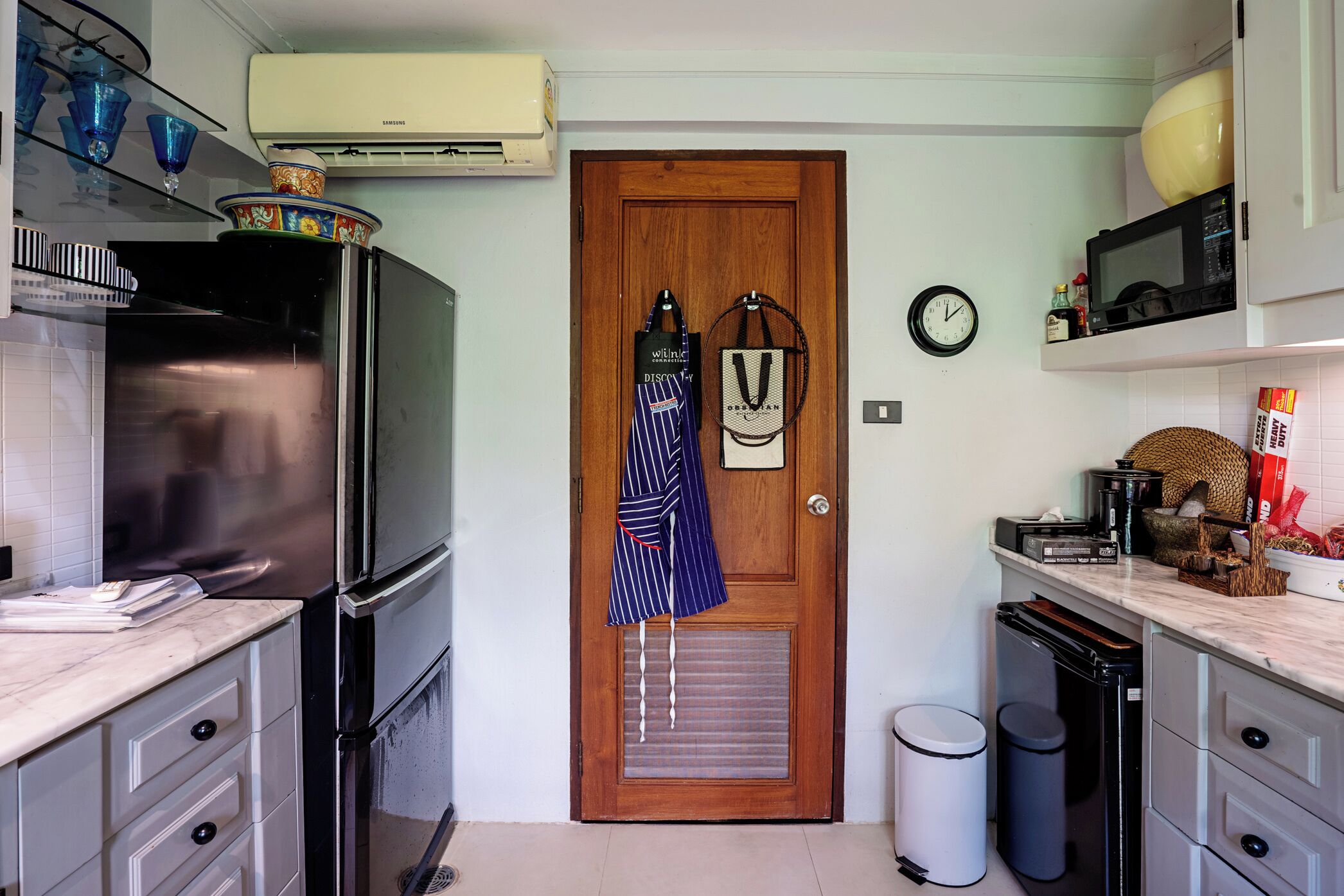 A compact kitchen with a refrigerator, microwave, and sink. There is a wooden door with a small window and a clock on the wall.