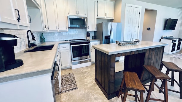 A modern kitchen with white cabinets, stainless steel appliances, and a central island with stools. - Near Chandelier of Gruene, Downtown & Whitewater – Game Room, Fire Pit & More! (New Braunfels)