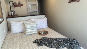 Un dormitorio con cama, almohadas, una colcha, una mesita de noche con lámpara, una planta pequeña y un cartel en la pared.