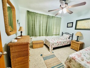 Une chambre à coucher comprenant un lit, deux tables de chevet, un ventilateur de plafond, un miroir et une fenêtre avec des rideaux.