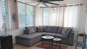 Ein Wohnzimmer mit einem Ecksofa, zwei kleinen runden Tischen, einem Deckenventilator und einem Fenster mit Jalousien.