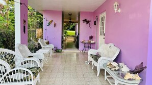 Ein überdachter Patio-Bereich mit weißen Korbmöbeln, einem kleinen Tisch mit Dekoration und Blick in einen Garten.