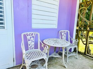 Une terrasse agrémentée de deux chaises blanches décorées et d’une petite table ronde, le tout adossé à un mur violet orné d’une fenêtre et d’une structure en treillis de bois.