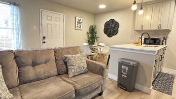 A living room with a sofa, a glass coffee table, a ceiling fan, and a kitchen area with cabinets and a microwave.