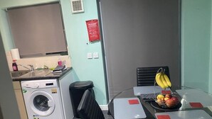 Une cuisine compacte équipée d’un lave-linge, d’un évier et d’une petite table avec un panier de fruits et une chaise.