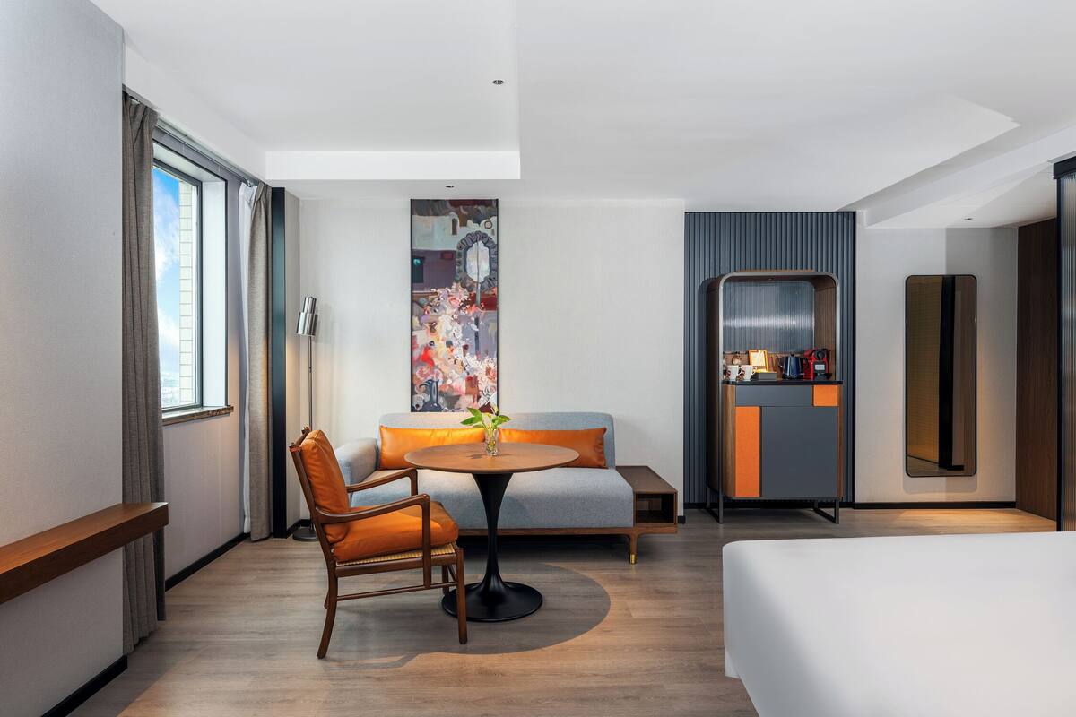 A modern hotel room with a bed, a round table with a vase, a sofa, a chair, a desk, and a large mirror.