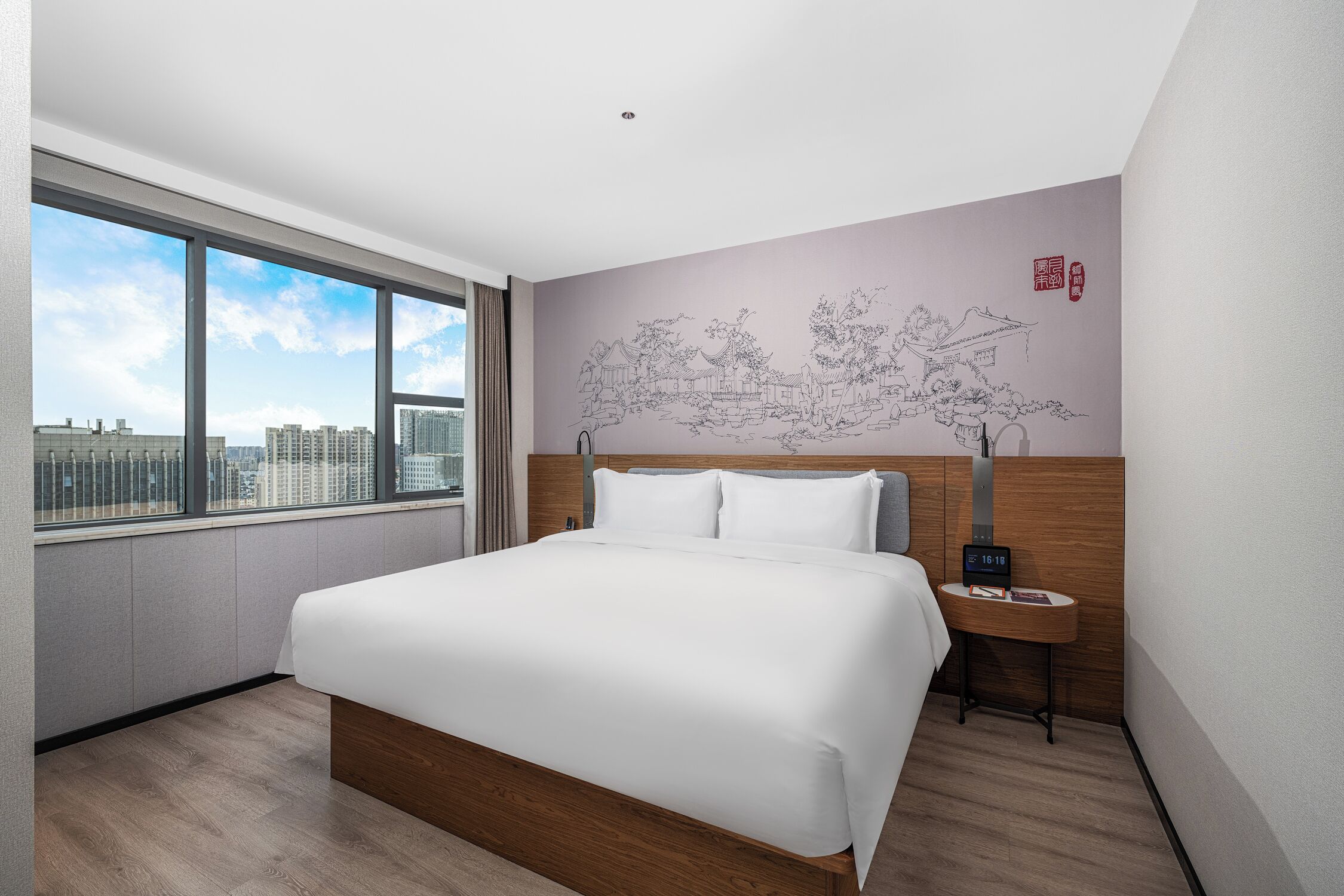 Una habitación de hotel moderna con una cama grande, una mesita de noche, un reloj en la pared y vistas de la ciudad a través de amplios ventanales.