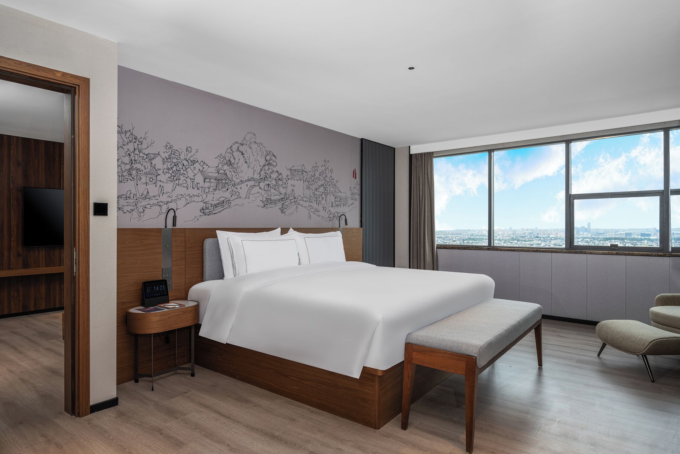 A modern hotel room with a large bed, a wooden nightstand, a bench, a TV, and a large window with a city view.