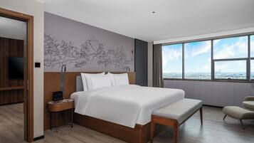 A modern hotel room with a large bed, a wooden nightstand, a bench, a TV, and a large window with a city view.