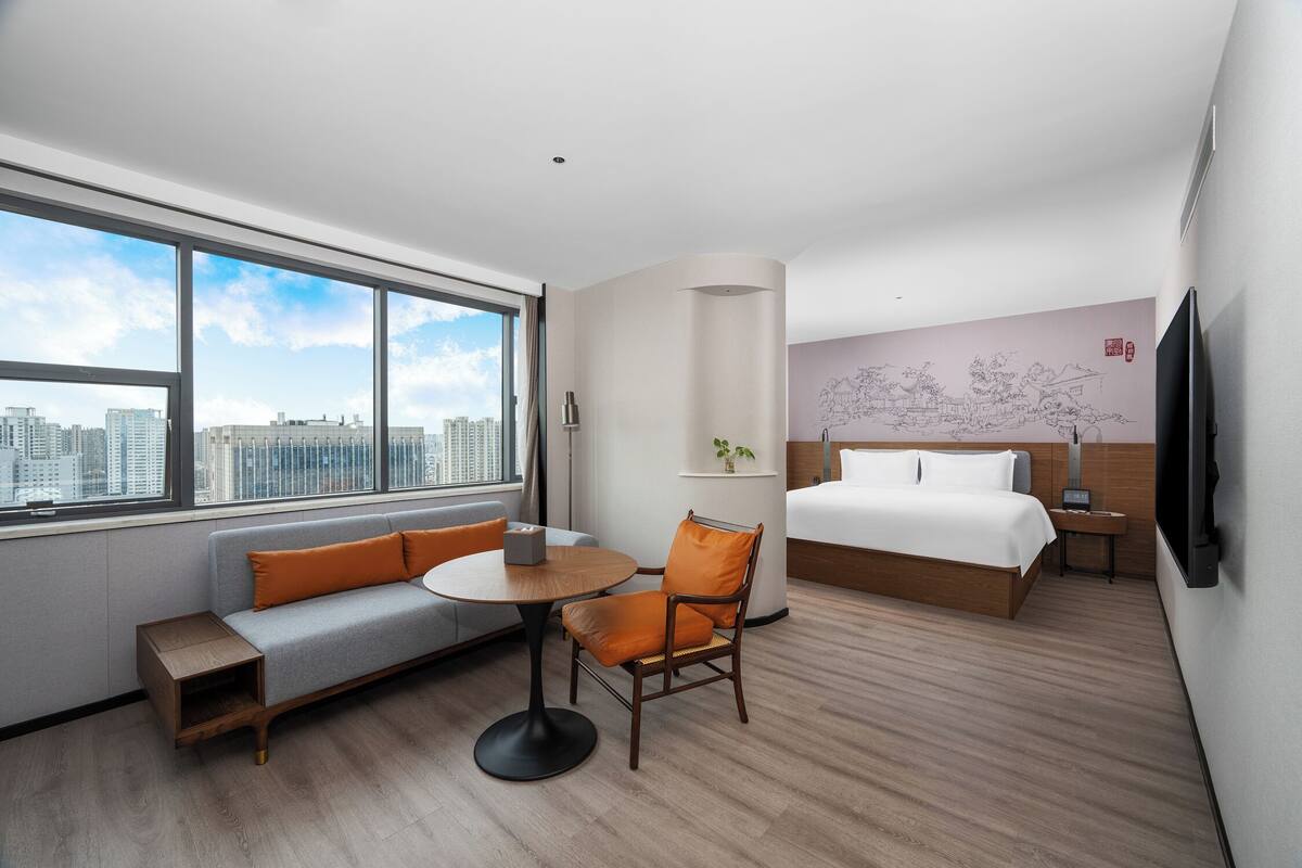 A hotel room with a large window, a sofa, a round table, a chair, a bed, a TV, and a wall with artwork.