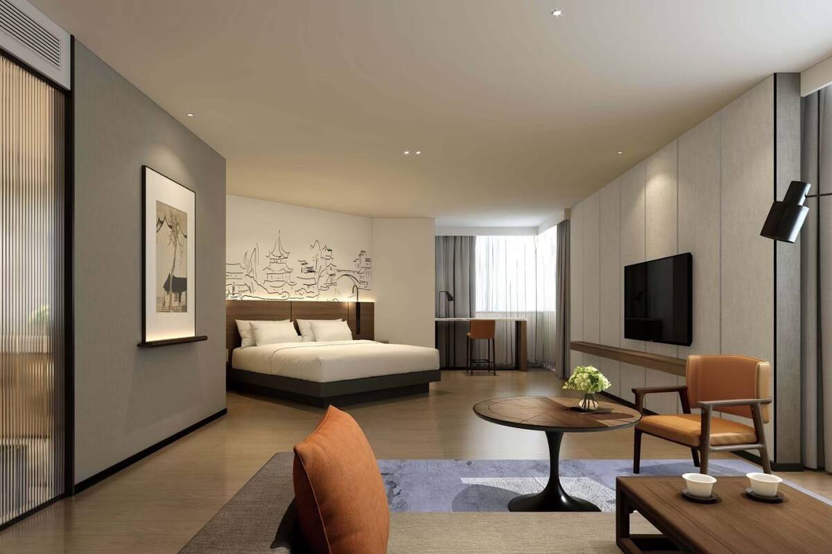 A modern hotel room with a bed, a desk, a TV, and a seating area with a table and chairs.