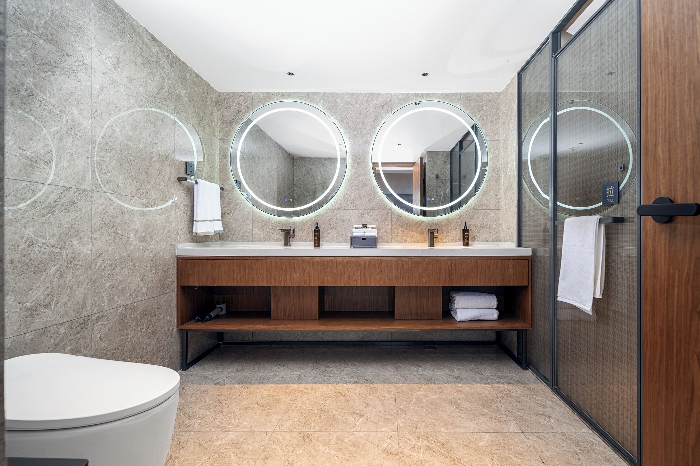 A modern bathroom with a marble wall, a wooden vanity, a toilet, and a glass shower enclosure.