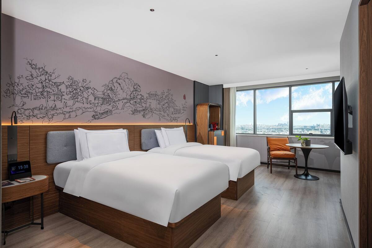 A modern hotel room with two beds, a small table, a chair, a TV, and a large window with a city view.