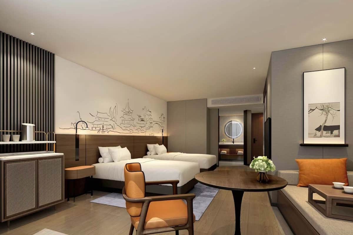 A modern hotel room with a large bed, a desk, a chair, and a small table. There is a wall-mounted shelf with cups and a decorative wall piece.