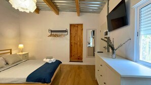 A bedroom with a bed, bedside table, lamp, wooden door, TV, and a window with blinds. - MarketHome Ecolodge Plataneras (Lomo Del Balo (Guia I.))