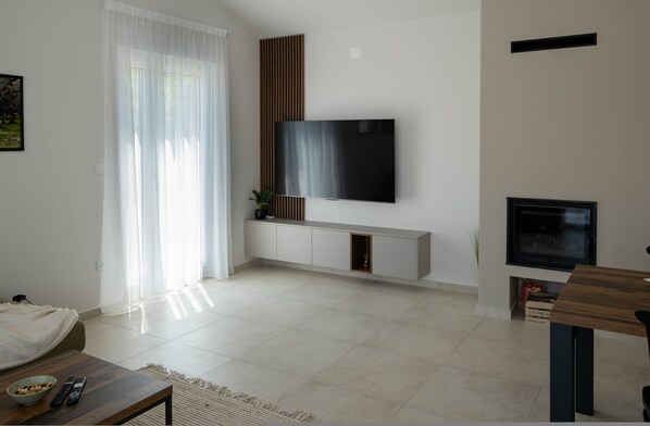 Ένα μοντέρνο σαλόνι με μια τηλεόραση επίπεδης οθόνης τοποθετημένη σε έναν λευκό τοίχο, ένα ξύλινο έπιπλο για την τηλεόραση, έναν καναπέ και ένα τραπέζι τραπεζαρίας με καρέκλες.